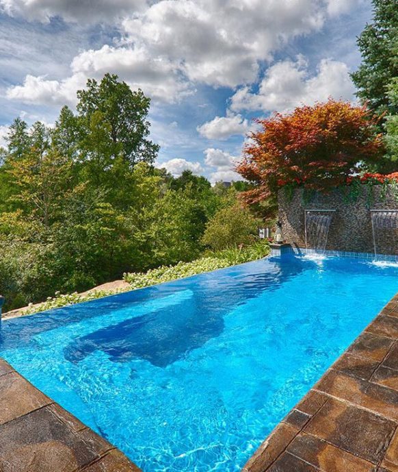 Infinity pool with clear blue water on a stone patio, overlooking a lush green landscape with trees and a partly cloudy sky. Water flows from two spouts on a stone wall, and the surrounding area has some outdoor seating.